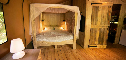 Vista interna della tenda Safari Lodge al Camping Les 3 Cantons, Francia, con letto a baldacchino e arredi in legno.