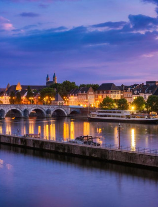 Vista nocturna de una ciudad iluminada junto al río cerca de Herkenbosch, Limburgo, Países Bajos, con puente antiguo.