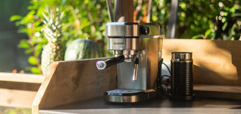 Cafetera espresso y espumador de leche sobre mesa de madera, con piña y melón al fondo, al aire libre.