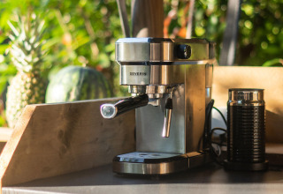Cafetera espresso y espumador de leche sobre mesa de madera, con piña y melón al fondo, al aire libre.