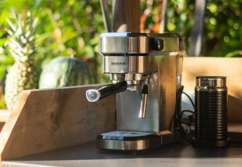 Cafetera espresso y espumador de leche sobre mesa de madera, con piña y melón al fondo, al aire libre.