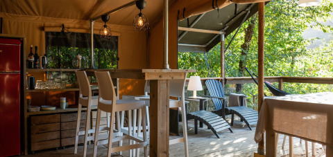Safari lodge tent interior at Camping Delle Rose, Italy, featuring bar stools, a veranda, and lush trees.