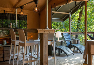 Safari lodge tent interior at Camping Delle Rose, Italy, featuring bar stools, a veranda, and lush trees.