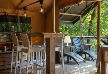 Safari lodge tent interior at Camping Delle Rose, Italy, featuring bar stools, a veranda, and lush trees.