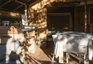 Interno di una tenda safari lodge con cucina, tavolo da pranzo e divano, sole e ombre, Camping Delle Rose, Italia.