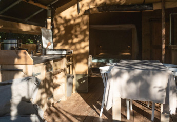 Interno di una tenda safari lodge con cucina, tavolo da pranzo e divano, sole e ombre, Camping Delle Rose, Italia.