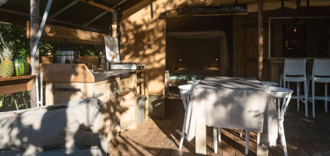 Interno di una tenda safari lodge con cucina, tavolo da pranzo e divano, sole e ombre, Camping Delle Rose, Italia.