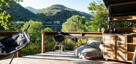 Terraza de madera en Safari Lodge con hamaca, puff, barbacoa y cocina exterior con vistas a colinas verdes.