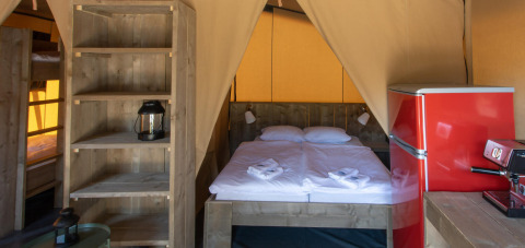 Interior de la tienda safari Base Lodge en Camping Delle Rose, Italia, con cama doble y nevera roja visible.