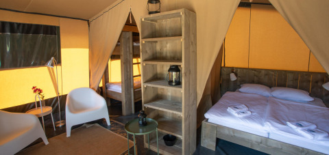 Interior de tienda safari Base Lodge en Camping Delle Rose, Italia, con cama, sillas y estantería.