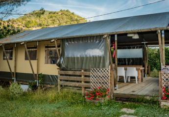 Tienda safari llamada Wood Lodge con terraza cubierta de madera, rodeada de colinas verdes y flores rojas.