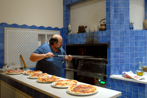 Hombre horneando pizzas en una cocina de azulejos azules en Camping Delle Rose, Liguria, Italia.