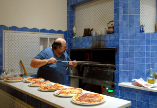 Uomo che prepara pizze in una cucina con piastrelle blu al Camping Delle Rose, Liguria, Italia.