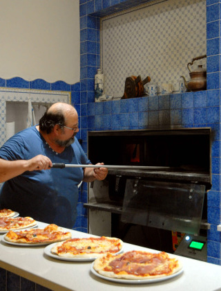 Hombre horneando pizzas en una cocina de azulejos azules en Camping Delle Rose, Liguria, Italia.