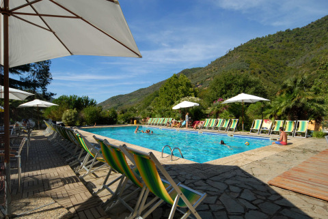 Piscina all’aperto con lettini e ombrelloni circondata da colline al Camping Delle Rose in Liguria, Italia.