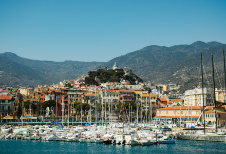 Boats docked in a marina with colorful buildings and mountains near Isolabona, Liguria, Italy.