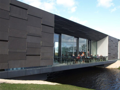Modern gebouw met glaswanden en bezoekers op een terras boven het water in Camping Vrijhaven Heeg.