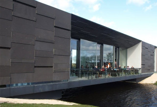 Moderne bygning med glasfacade og folk, der sidder på en terrasse over vandet i Camping Vrijhaven Heeg.