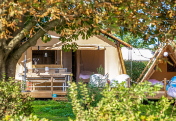 Safari-teltet Cozy Lodge + sanitetsfaciliteter omgivet af grønne planter og træer på en solrig dag.