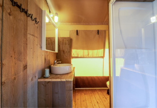 Intérieur d’une tente safari Cozy Lodge avec salle de bain en bois, lavabo, miroir et douce lumière.