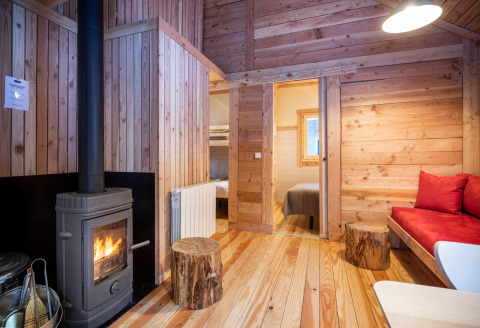Knusse glampinghut met houtkachel, houten wanden en zicht op woonruimte en slaapkamer vanbinnen.