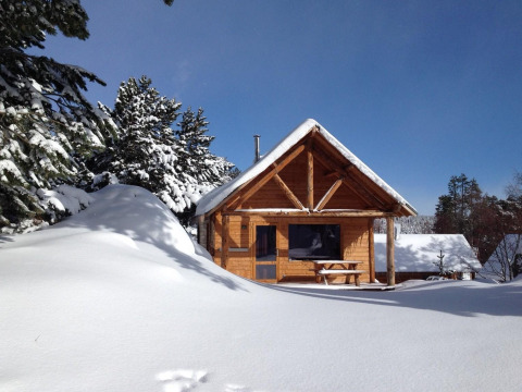 Cabaña de madera cubierta de nieve, rodeada de pinos, bajo un cielo azul claro y soleado.