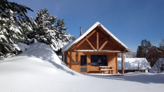 Cabaña de madera cubierta de nieve, rodeada de pinos, bajo un cielo azul claro y soleado.
