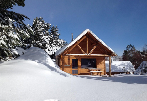 Cabaña de madera cubierta de nieve, rodeada de pinos, bajo un cielo azul claro y soleado.
