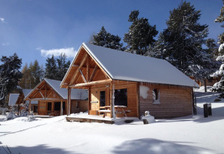Besneeuwde houten chalets bij Les Chalets Huttopia de Font-Romeu in Frankrijk, tussen winterse bomen.