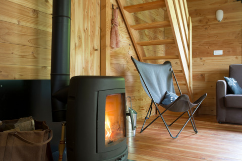 Intérieur de chalet en bois avec poêle à bois, fauteuil moderne et escalier, Les Chalets Huttopia Font-Romeu.