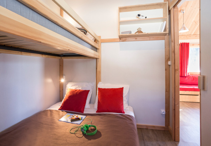 Knusse slaapkamer in een chalet met houten stapelbed, tweepersoonsbed, rode kussens en zicht op leefruimte.