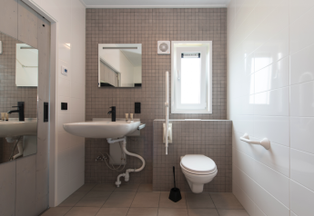 Baño accesible con inodoro suspendido, lavamanos, barras de apoyo y espejo en Access Lodge, Kompas Camping.