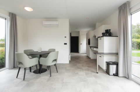 Moderne lodge interiør med spisebord, fire stole, køkkenområde og store vinduer på Kompas Camping - Westende.