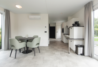 Interior de lodge moderno con mesa redonda, cuatro sillas, área de cocina y grandes ventanas en Kompas Camping.