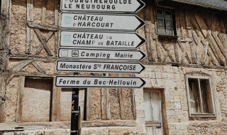 Señales direccionales frente a una casa de entramado de madera en un pueblo francés, cielo azul arriba.