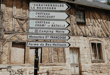 Señales direccionales frente a una casa de entramado de madera en un pueblo francés, cielo azul arriba.