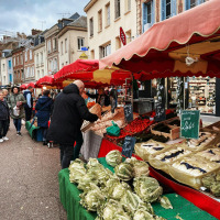 Escena de mercado en Moyaux, Normandía, Francia, con coloridos puestos de verduras y gente comprando productos frescos.