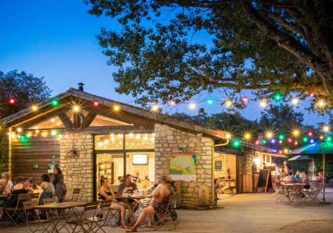 Zona de comedor al aire libre con luces y gente cenando en Huttopia le Moulin, Auvergne-Rhône-Alpes, Francia.