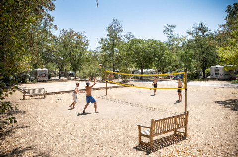People play volleyball at a campsite in Huttopia le Moulin, surrounded by trees, benches, and camper vans.