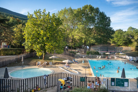 Piscina al aire libre en Huttopia Divonne Les Bains con familias disfrutando y relajándose bajo los árboles.