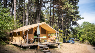 Dos personas descansando frente a una tienda safari Toile&bois tent Classic en un camping rodeado de árboles altos.