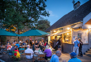 Outdoor dining area at Huttopia Divonne Les Bains with people enjoying a summer evening under green umbrellas.