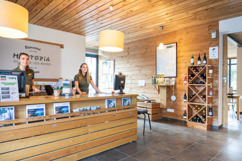 Recepción de Huttopia Divonne Les Bains con dos empleadas, paredes de madera y estante de vinos en Francia.