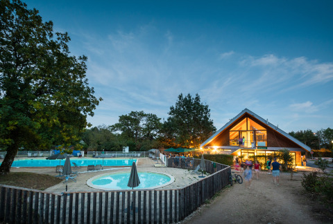 Avondbeeld van Huttopia Divonne Les Bains vakantiepark met zwembad en verlicht hoofdgebouw in Frankrijk.