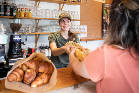 Panadera sonriente entrega pan recién hecho a cliente en Huttopia Divonne Les Bains, parque vacacional.