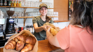Panadera sonriente entrega pan recién hecho a cliente en Huttopia Divonne Les Bains, parque vacacional.