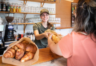Smiling baker hands fresh bread to customer at bakery counter in Huttopia Divonne Les Bains holiday park.