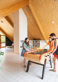 Sala interior en Huttopia Divonne Les Bains, Francia, con adultos jugando futbolín y relajándose.
