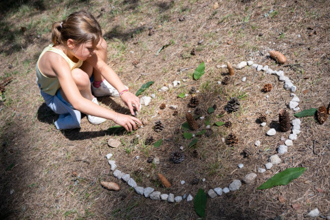 Niña crea arte con piñas, piedras y hojas en el suelo del parque Huttopia Divonne Les Bains en Francia.