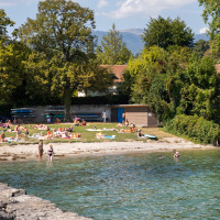 Personas descansan y nadan en una playa de lago cerca de Divonne Les Bains, rodeados de naturaleza verde.